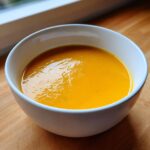 A close-up of vibrant orange, creamy butternut squash soup served in a white bowl on a wooden surface.