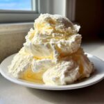 A large mound of creamy, pale yellow cottage cheese ice cream served on a white plate near a sunlit window.