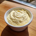 A small white bowl filled with thick, creamy, yellow garlic aioli, sitting on a wooden surface.