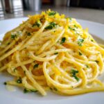 A close-up of a serving of creamy lemon pasta, garnished generously with bright yellow lemon zest and chopped green parsley.