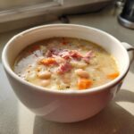 Close-up of a white mug filled with creamy navy bean soup, topped with shredded ham and diced carrots.