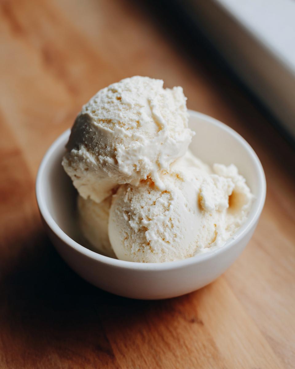 Two scoops of creamy, vanilla-colored protein ice cream served in a small white bowl on a wooden surface.