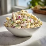 A close-up of a white bowl filled with creamy tuna pasta salad featuring rotini pasta, red onion, and celery.