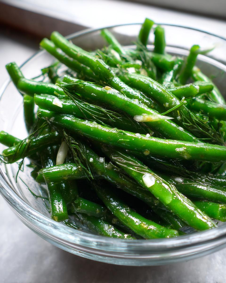 Close-up of vibrant green dilly beans mixed with fresh dill and garlic pieces in a clear glass bowl.