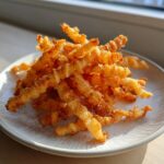 A close-up of a pile of crispy, golden, curly cheese straws seasoned and served on a light-colored plate.