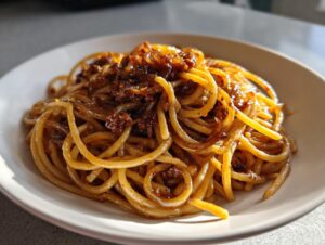 Close-up of glossy spaghetti tossed with dark, rich caramelized onion pasta sauce in a light bowl.