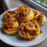 Close-up of five freshly baked sausage pinwheels topped with melted cheese on a white plate.