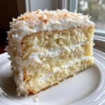 A tall slice of fluffy coconut cake recipe showing layers of white cake and frosting, topped with toasted coconut flakes.