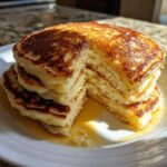 A tall stack of fluffy discard pancakes, with a piece cut out, drizzled generously with maple syrup.