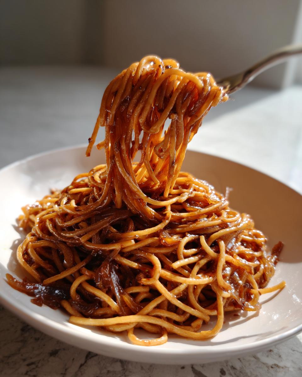 A fork lifts a generous twirl of glossy caramelized onion pasta from a white bowl.