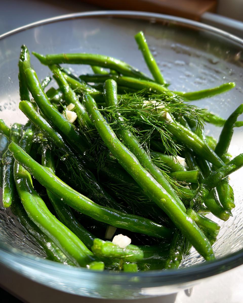 Close-up of bright green dilly beans mixed with fresh dill fronds and chopped garlic in a clear glass bowl.