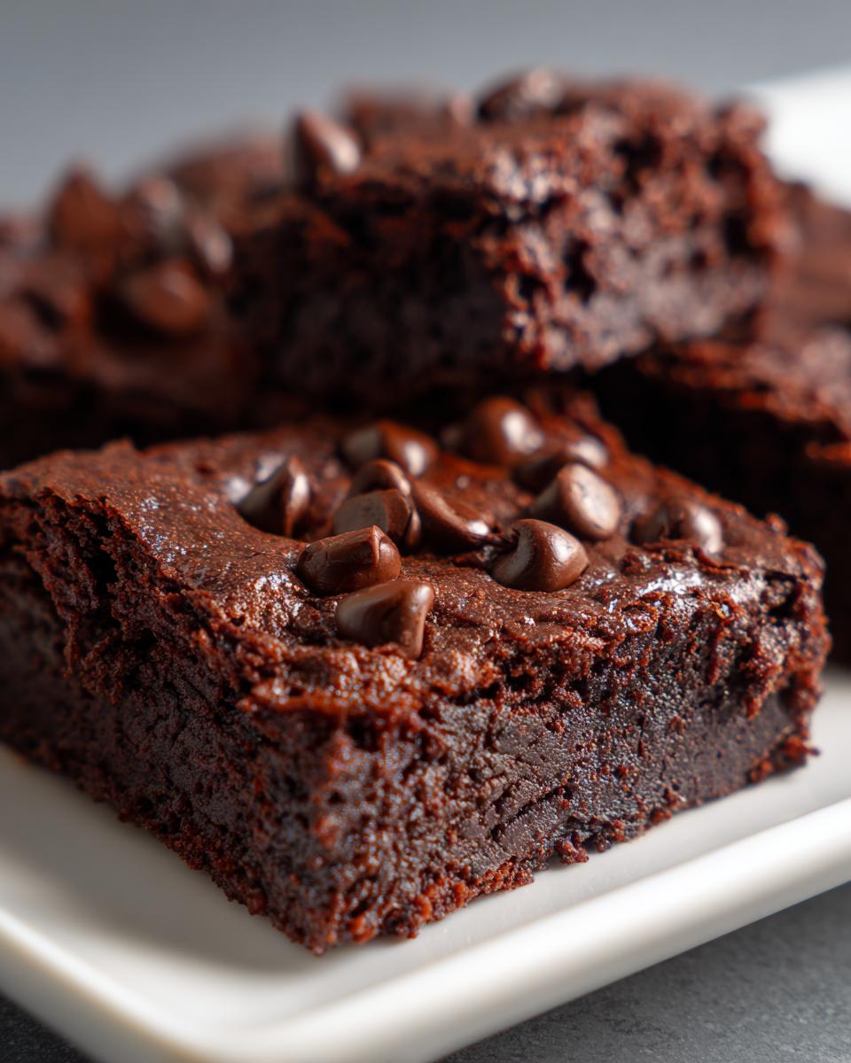 Close-up of a rich, fudgy black bean brownie square topped with melted chocolate chips on a white plate.