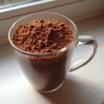 A clear glass mug filled to the brim with rich brown homemade hot cocoa mix powder.
