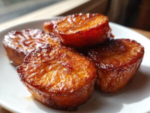 Four thick slices of candied sweet potatoes glistening with a rich, dark glaze on a white plate.