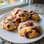 Four freshly baked cranberry orange cookies drizzled with white glaze, resting on a white plate near a window.