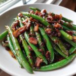 Close-up of bright green beans tossed in a savory glaze and topped with crispy bacon pieces.