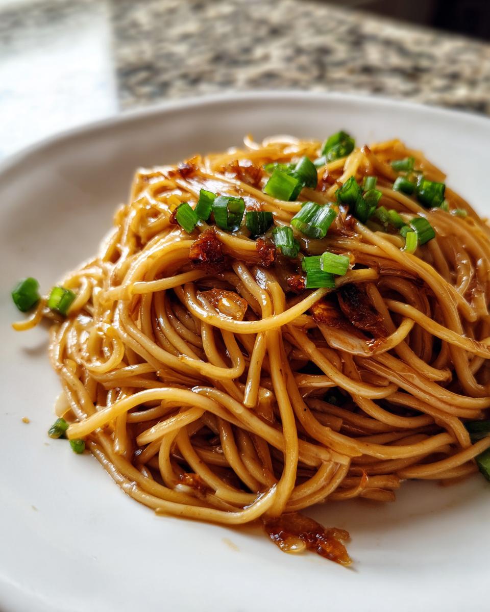 A close-up of a serving of glossy, saucy garlic noodles topped with chopped green onions.