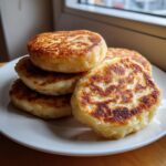 A stack of freshly cooked, golden brown Irish potato farls resting on a white plate near a window.