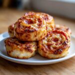 A close-up of several golden brown, baked pizza pinwheels stacked on a white plate.