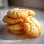 A close-up stack of golden, slightly cracked butter cookie treats dusted with sugar, sitting on a white marble surface.