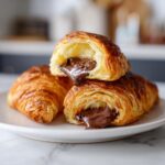 Close-up of three flaky chocolate croissants, one cut open revealing melted chocolate filling.