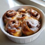 Close-up of several gooey cinnamon roll bites topped with white icing, served in a small white bowl.