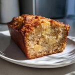 A close-up of a slice of golden brown Hawaiian banana bread, showing moist crumb and chunks of pineapple or nuts.