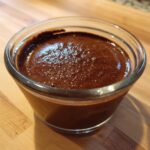 A close-up view of rich, dark homemade chocolate pudding served in a small clear glass cup on a wooden surface.