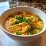 A close-up of a white bowl filled with hot egg drop soup, featuring wispy egg ribbons and green onions.