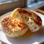 Two halves of a homemade english muffin, toasted golden brown, showing the signature nooks and crannies, topped with melting butter.