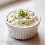 Close-up of creamy, chunky tartar sauce recipe garnished with fresh dill in a white fluted ramekin.