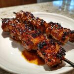 Close-up of three perfectly grilled honey garlic chicken skewers with a dark, sticky glaze on a white plate.