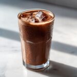 A tall glass of iced chocolate milk covered in condensation, sitting on a bright marble surface.