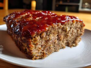 Close-up of a thick slice of juicy meatloaf topped with a shiny, reddish-brown glaze, served on a white plate.