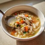 A spoonful of hearty turkey soup showing shredded turkey, pearl pasta, carrots, and celery in a white bowl.
