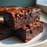 Close-up of stacked, fudgy black bean brownies topped with melted chocolate chips on a white plate.