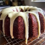 A close-up of a rich, moist red velvet bundt cake drizzled heavily with white cream cheese glaze.