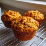 Three golden-brown morning glory muffins cooling on a wire rack with visible shredded carrots.