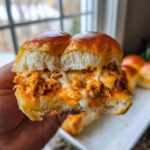 Close-up of a hand holding a delicious buffalo chicken sliders, showing shredded chicken coated in sauce and gooey melted cheese.