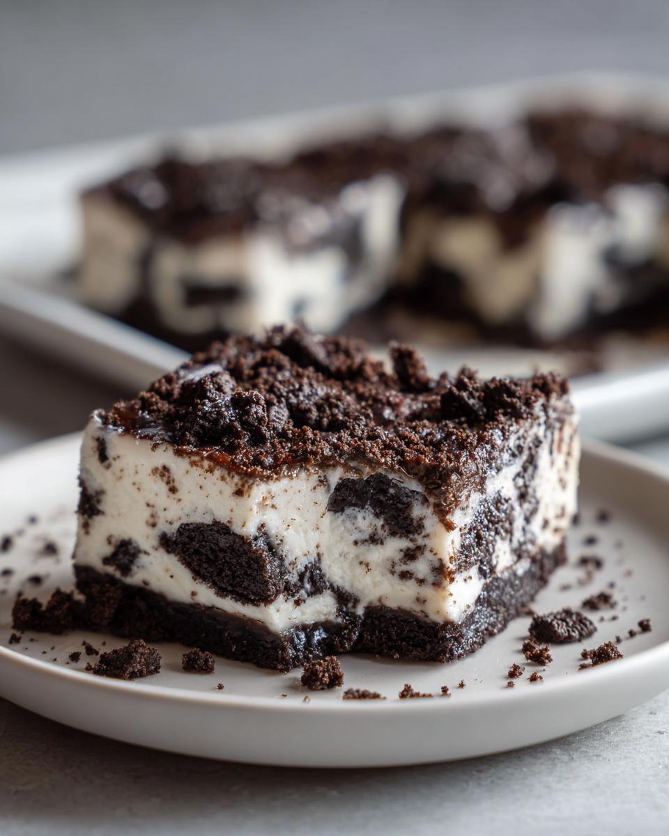 A decadent square slice of Oreo fudge with a creamy white filling mixed with dark cookie pieces, topped with crushed Oreo crumbs.