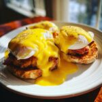 Close-up of two servings of eggs benedict, featuring poached eggs, hollandaise sauce, and bacon on toasted English muffins.