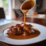 A white ladle pours rich, steaming chicken gravy over pieces of seasoned meat on a white plate.
