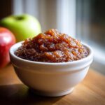A close-up of thick, dark brown homemade apple butter served in a small white bowl, with fresh apples in the background.