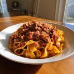 A close-up of fresh tagliatelle pasta generously topped with rich, slow-cooked bolognese sauce in a white bowl.