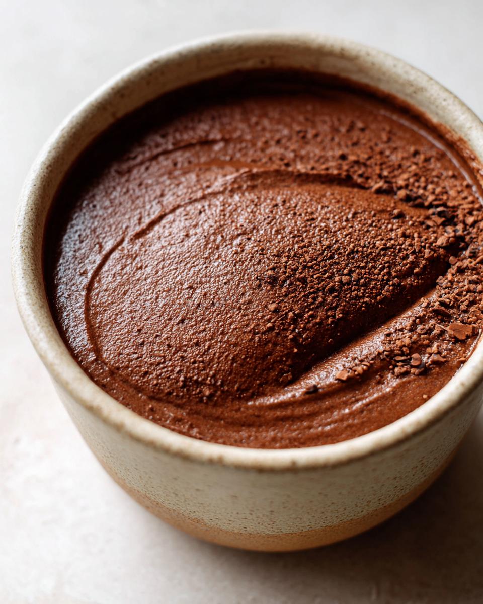Close-up of rich, dark chocolate pudding topped with cocoa powder in a speckled ceramic bowl.
