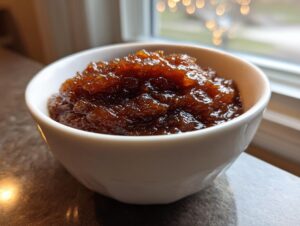 Close-up of thick, dark brown homemade apple butter served in a small white bowl near a window.