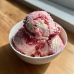 Three scoops of homemade strawberry ice cream with visible red swirls in a white bowl.