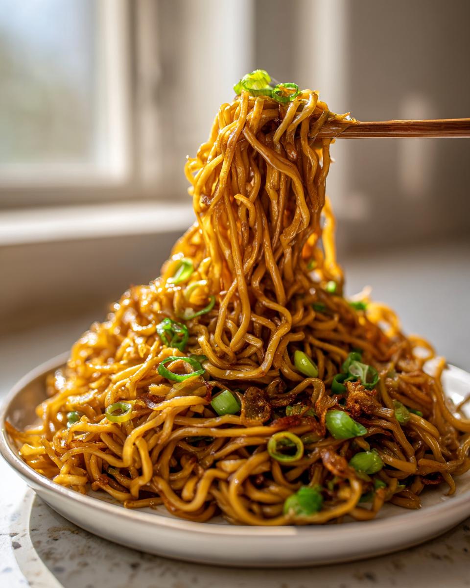 Close-up of a large serving of glossy, saucy garlic noodles being lifted by chopsticks, garnished with green onions.