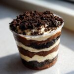 Close-up of a layered single serving dirt cake in a clear plastic cup, topped with cookie crumbs.