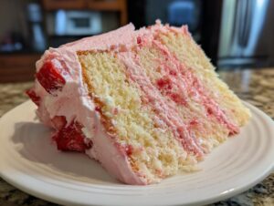 Close-up of a moist slice of strawberry cake with layers of vanilla sponge and pink strawberry filling/frosting.